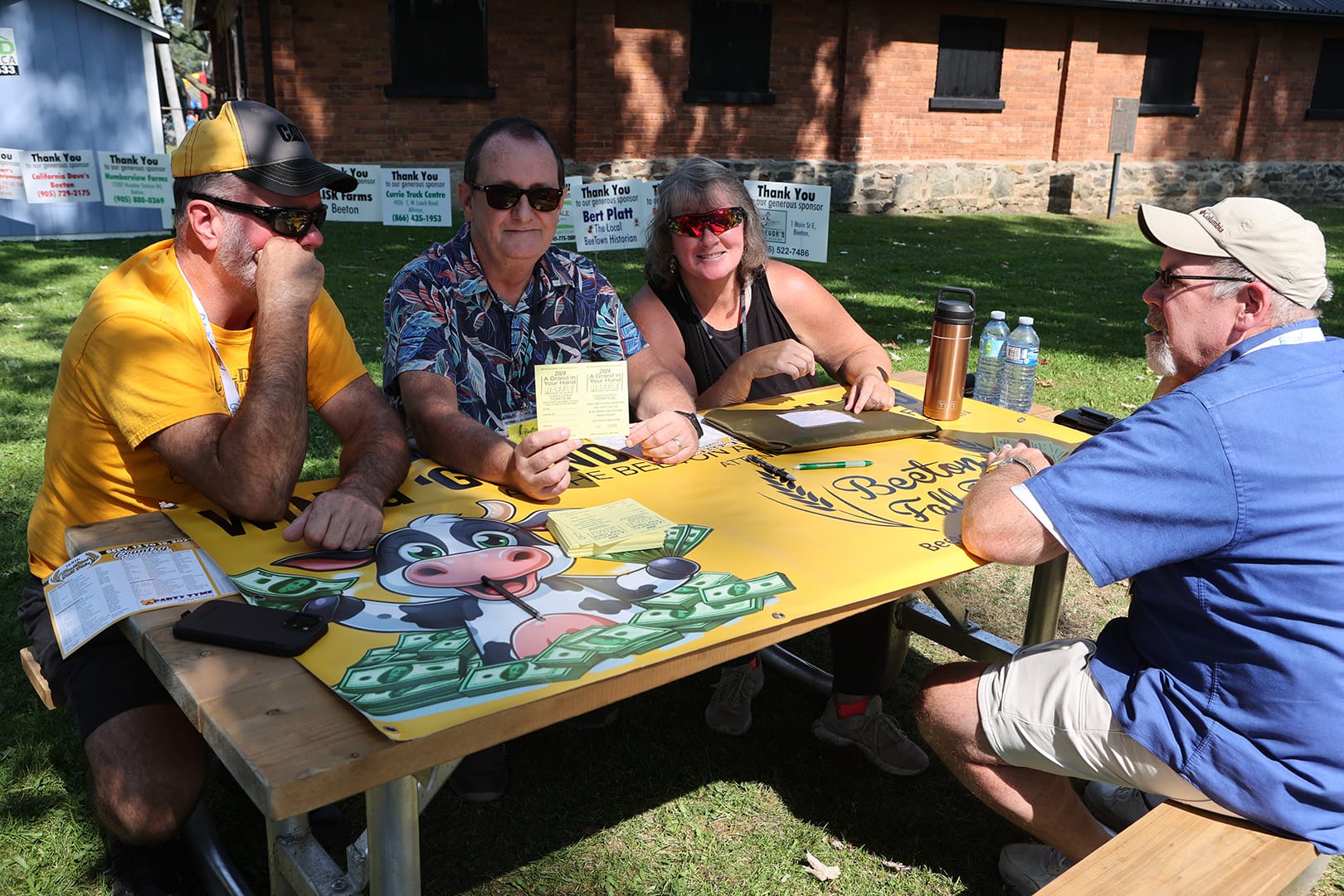 Four people sitting around a table with Beeton Fall Fair cow patty bingo game board and raffle tickets, representing community participation in fair games and raffles