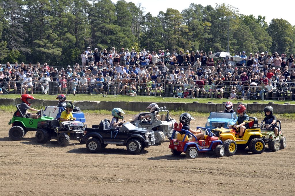 Children racing Power Wheels vehicles in derby competition at Beeton Fall Fair with crowd of spectators in bleachers watching the action