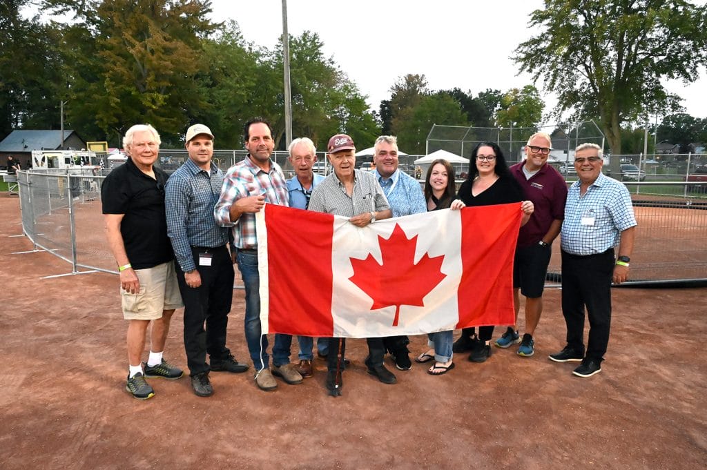 Group of Beeton Fall Fair dignitaries and board members holding Canadian flag at 2024 opening ceremonies on dirt track with fair infrastructure in background